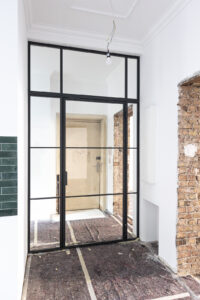 Stahl-Meister Glastrennwand mit Oberlicht, feststehenden Seitenteil und Drehtür, Raumtrenner zwischen Flur, Küche und Wohnzimmer aus Glas in Schwarz, transparentes VSG-Sicherheitsglas, guter Schallschutz, Einbau und Montage in Berlin