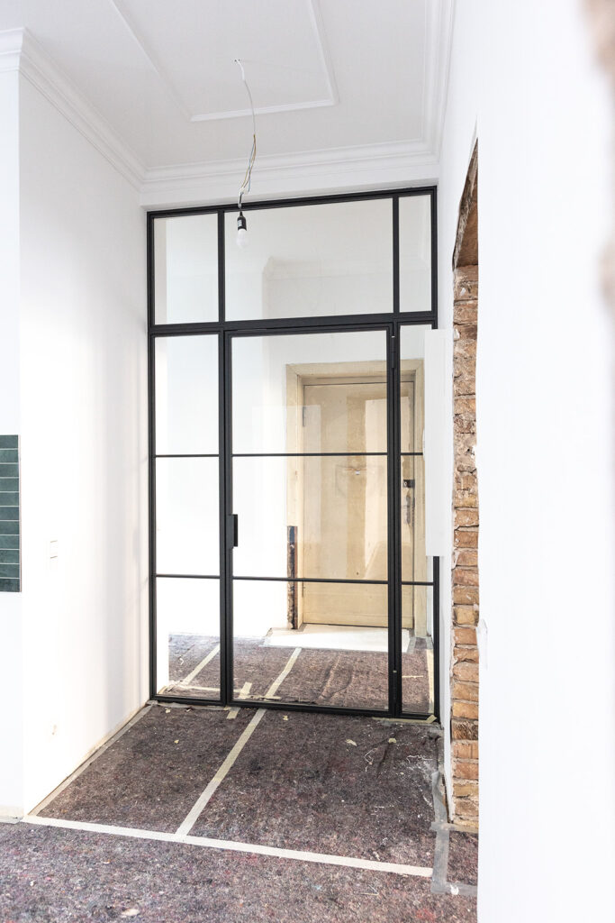 Stahl-Meister Glastrennwand mit Oberlicht, feststehenden Seitenteil und Drehtür, Raumtrenner zwischen Flur, Küche und Wohnzimmer aus Glas in Schwarz, transparentes VSG-Sicherheitsglas, guter Schallschutz, Einbau und Montage in Berlin