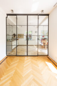 Stahl Meister Glastrennwand als Doppel-Schiebetür mit feststehenden Seitenteilen, VSG-Sicherheitsglas für guten Schallschutz, guter Wärmeschutz als Windfang für Küche und Wohnzimmer, Deckenhohe Lofttür aus Stahl und Glas, Aufmaß und Montage, Einbau in Berlin