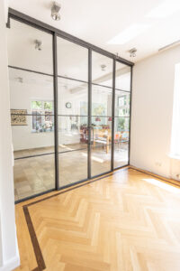 Stahl Meister Glastrennwand als Doppel-Schiebetür mit feststehenden Seitenteilen, VSG-Sicherheitsglas für guten Schallschutz, guter Wärmeschutz als Windfang für Küche und Wohnzimmer, Deckenhohe Lofttür aus Stahl und Glas