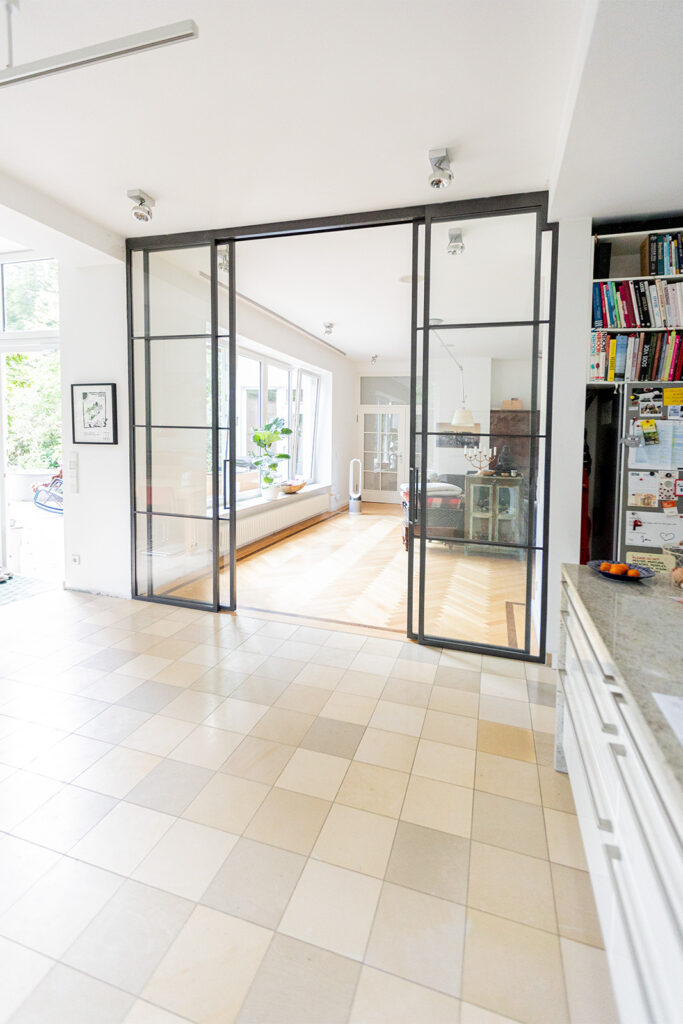 Stahl Meister Glastrennwand als Doppel-Schiebetür mit feststehenden Seitenteilen, VSG-Sicherheitsglas für guten Schallschutz, guter Wärmeschutz als Windfang für Küche und Wohnzimmer, Deckenhohe Lofttür aus Stahl und Glas